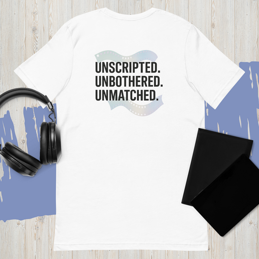 White unisex t-shirt laid on wood surface with bold black text reading "Unscripted. Unbothered. Unmatched." — styled with black headphones